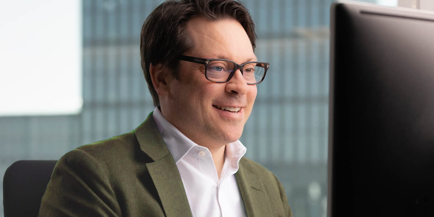 Mann mit Brille lächelt, während er vor einem Computer sitzt, im Hintergrund sind Gebäude.