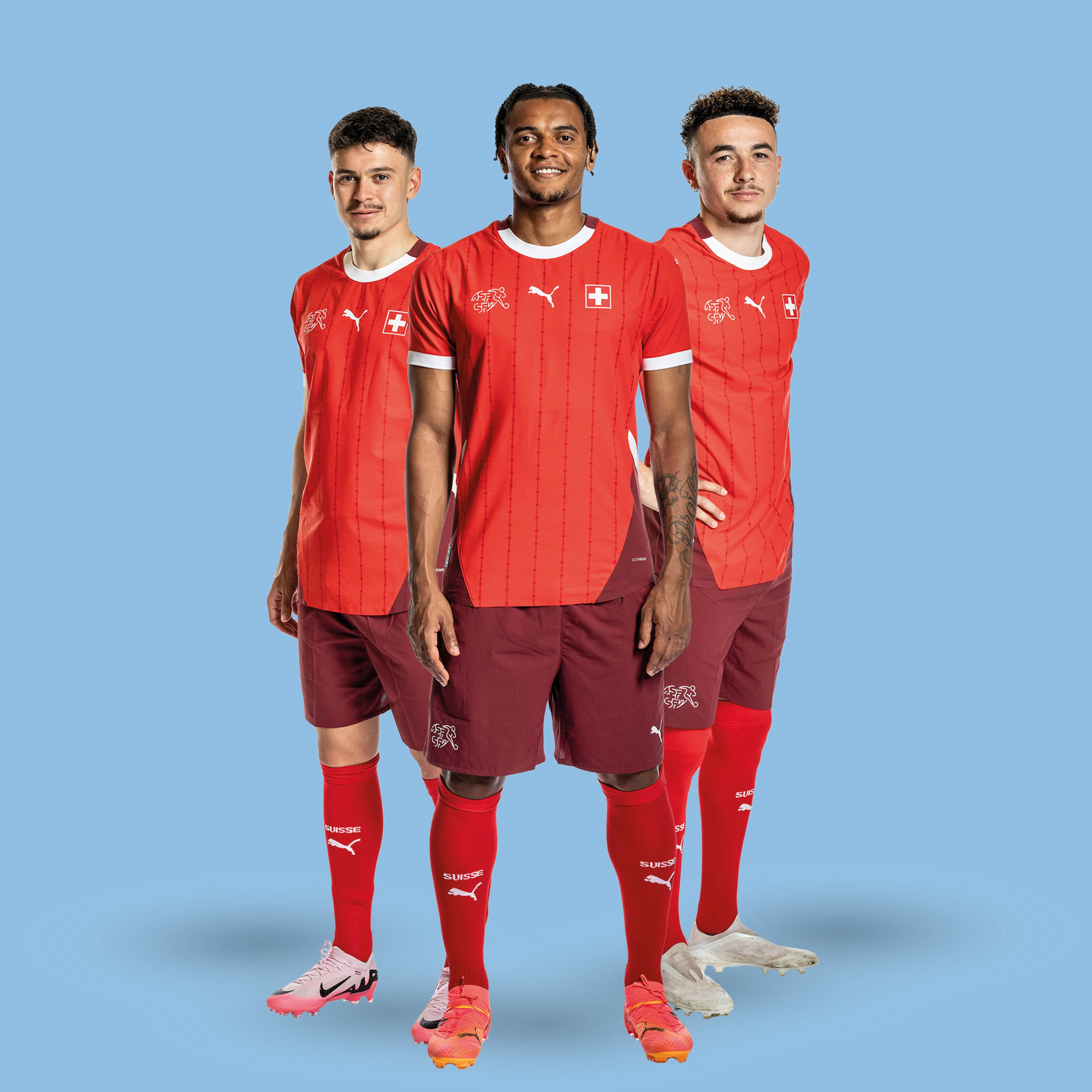 Drei Fußballspieler in roten Trikots stehen vor blauer Wand.