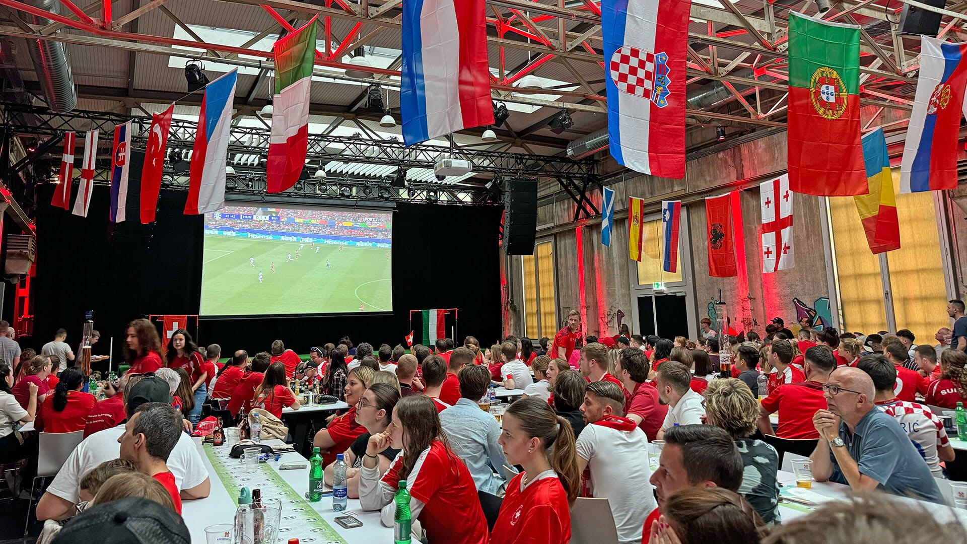 Une grande foule dans une salle regarde un écran de football. Beaucoup portent des vêtements rouges et plusieurs drapeaux sont accrochés.