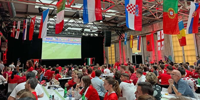 Une grande foule dans une salle regarde un écran de football. Beaucoup portent des vêtements rouges et plusieurs drapeaux sont accrochés. Une grande foule dans une salle regarde un écran de football. Beaucoup portent des vêtements rouges et plusieurs drapeaux sont accrochés.