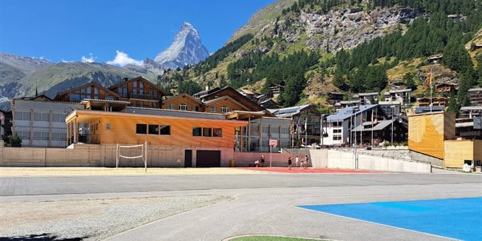 A landscape view featuring buildings, a sports area, and a mountain in the background. The sky is clear and blue, suggesting good weather. A landscape view featuring buildings, a sports area, and a mountain in the background. The sky is clear and blue, suggesting good weather.