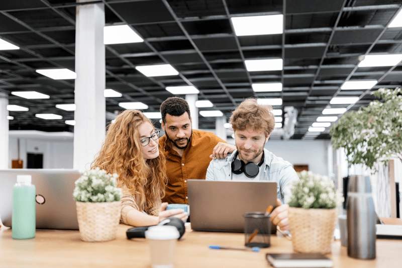 Das Bild zeigt drei junge Menschen, die gemeinsam an einem Tisch in einem modernen, offenen Büro arbeiten. Sie sitzen vor Laptops und sind von Pflanzen und Getränken umgeben. Die Atmosphäre wirkt entspannt und kollegial, als ob sie an einem Projekt zusammenarbeiten oder gemeinsam Ideen austauschen.