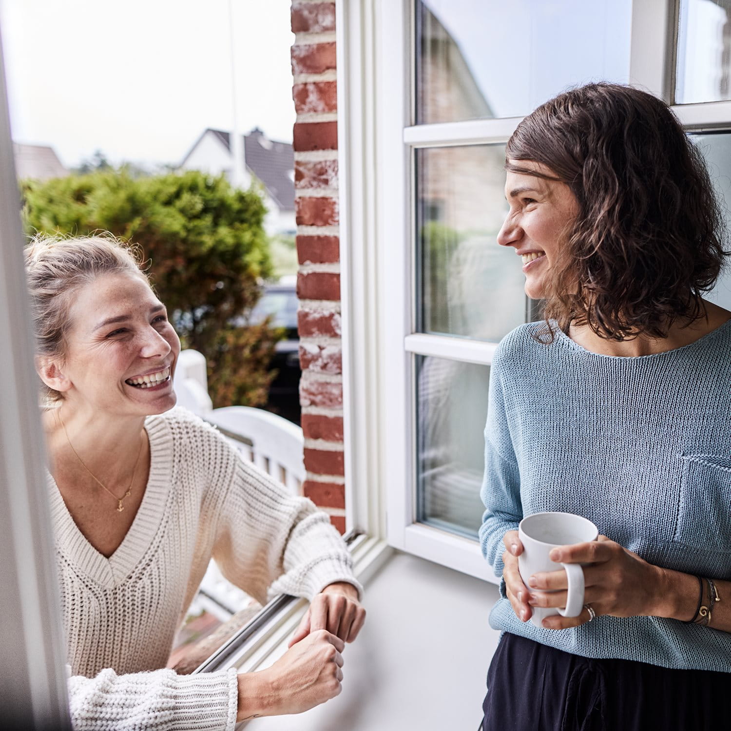 Zwei Frauen unterhalten sich entspannt am offenen Fenster eines Hauses. Eine Frau steht draußen und lehnt sich auf das Fensterbrett, die andere steht drinnen und hält eine Tasse Kaffee. Im Hintergrund sind ein Garten und Nachbarhäuser zu sehen.