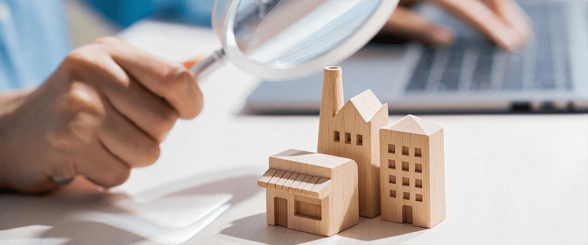Une main tient une loupe au-dessus de maquettes en bois de bâtiments sur une table.