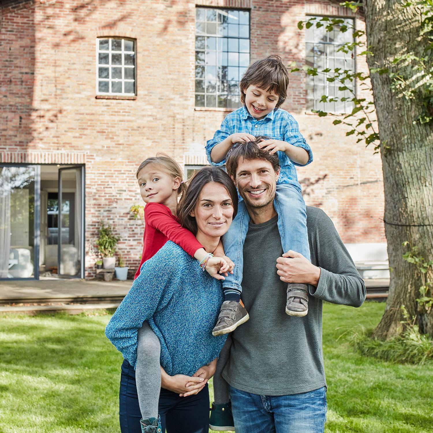 A family stands together in a garden in front of a brick house. One child is sitting on the father's shoulders, another is being carried by the mother.