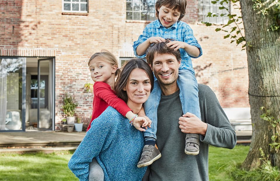 A family stands together in a garden in front of a brick house. One child is sitting on the father's shoulders, another is being carried by the mother. A family stands together in a garden in front of a brick house. One child is sitting on the father's shoulders, another is being carried by the mother.