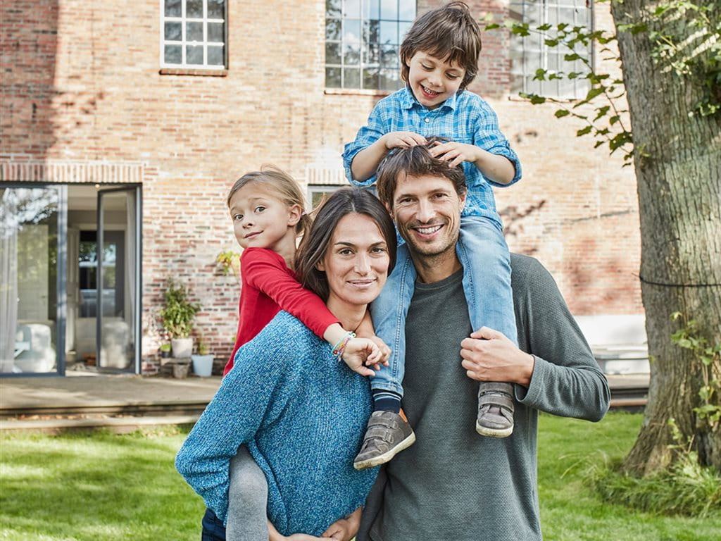 A family stands together in a garden in front of a brick house. One child is sitting on the father's shoulders, another is being carried by the mother.