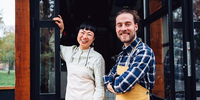 Two people wearing aprons standing at the entrance of a shop or café.
