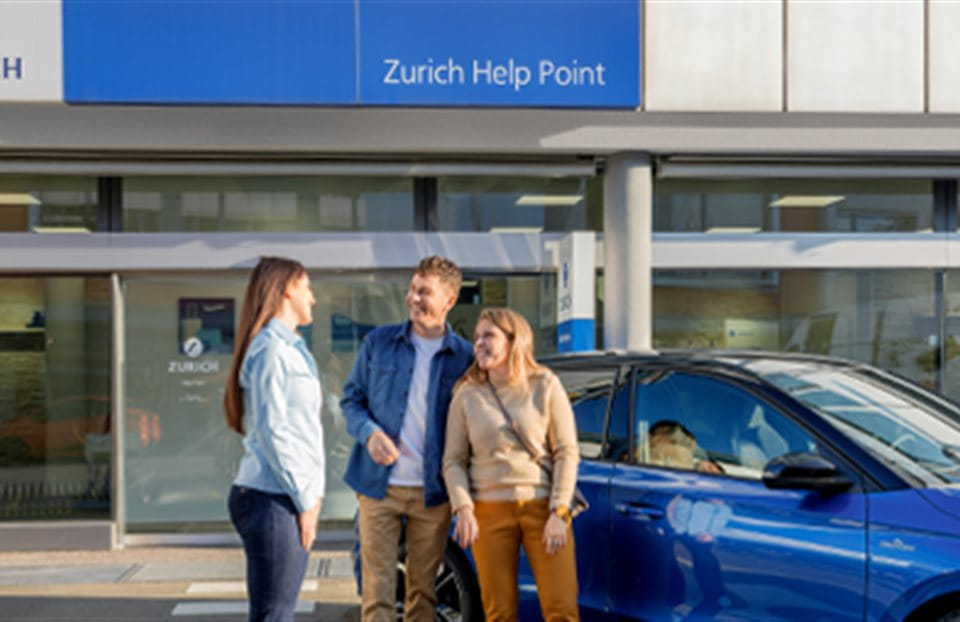 Three people are standing in front of a blue car and talking outside a Zurich Help Point. Three people are standing in front of a blue car and talking outside a Zurich Help Point.